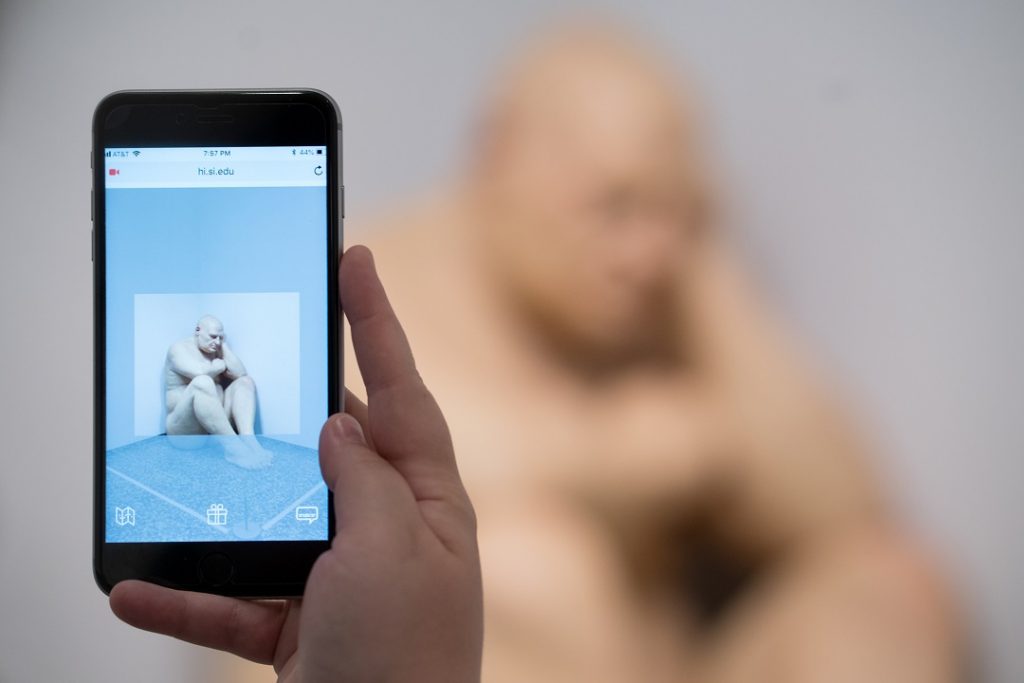 A hand uses a mobile phone, with a camera feature pulling up the image of a figurative sculpture behind it.