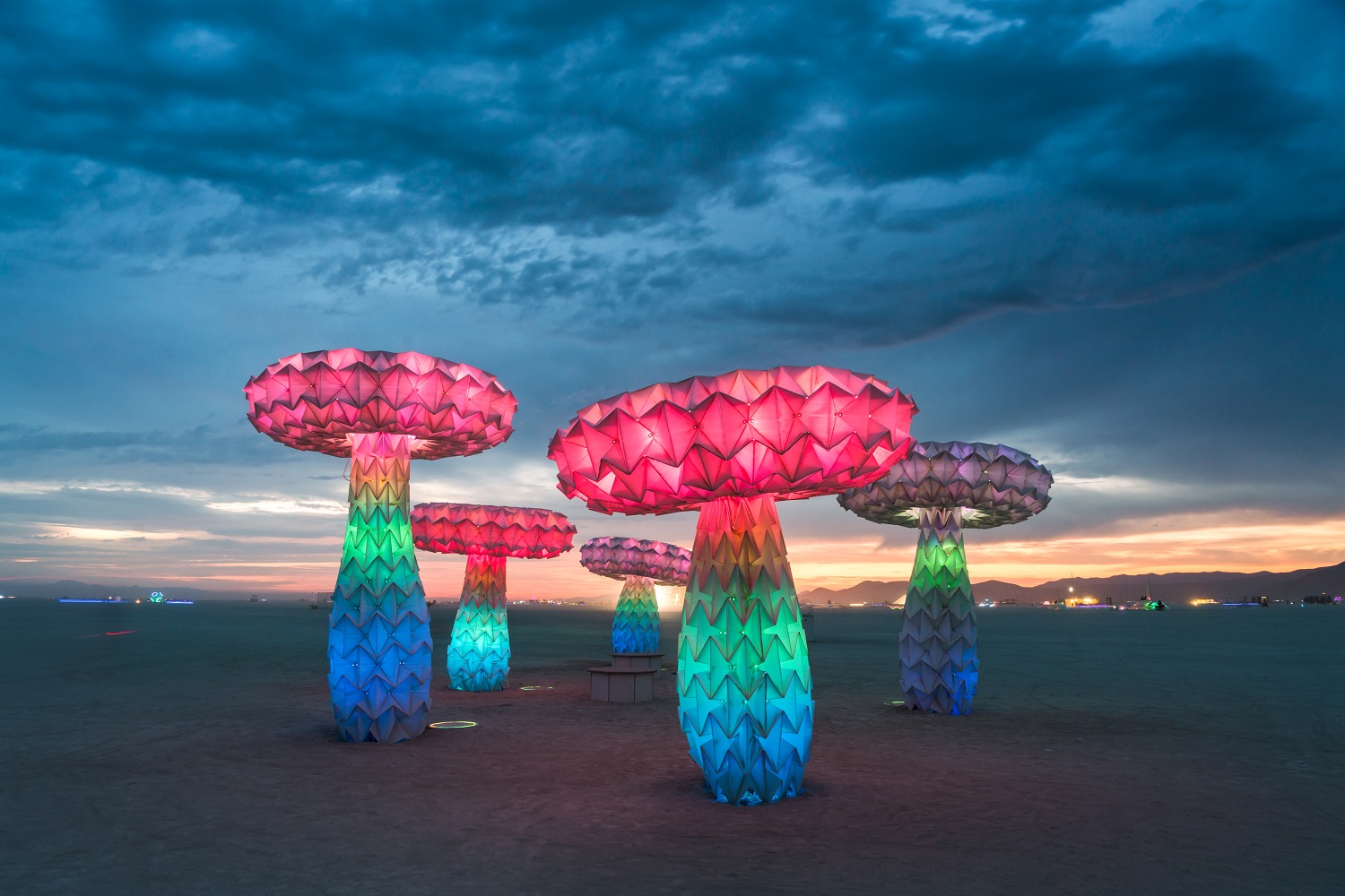 Pink, green, and blue lighted mushrooms against a dark, cloudy sky in a flat desert landscape.